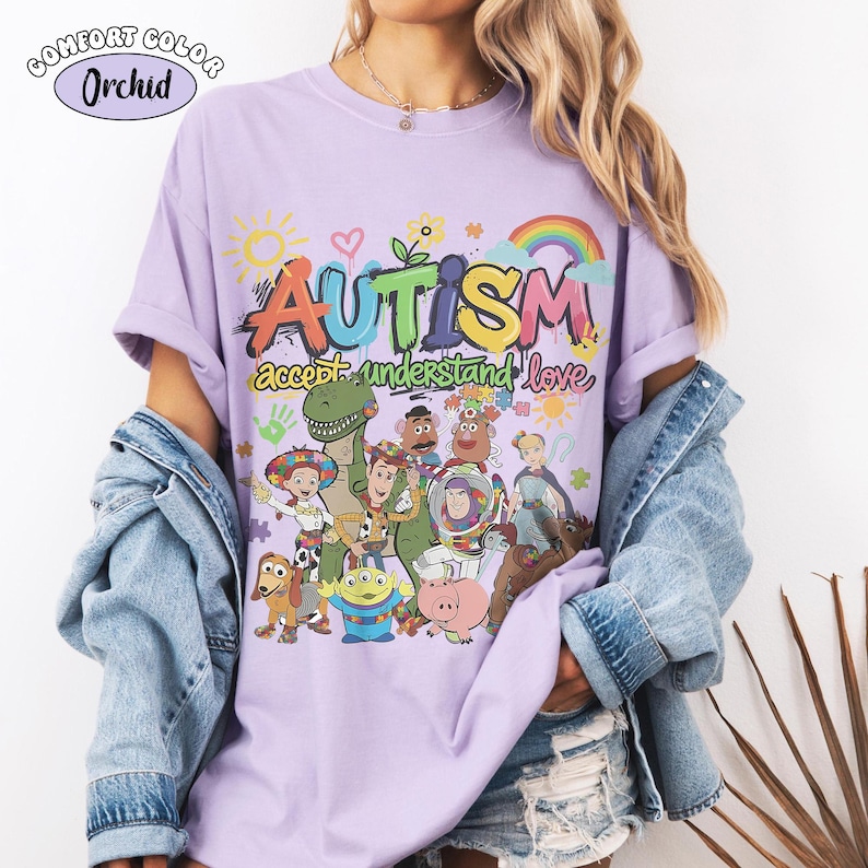 May include: A light purple t-shirt featuring a colourful graphic with the text "AUTISM accept understand love" and various cartoon characters including a cowboy, space ranger, dinosaur, and pig. The design includes a sun, rainbow, and puzzle pieces.