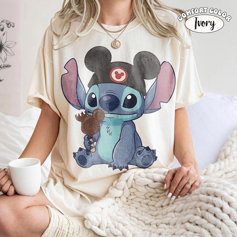 May include: An ivory-colored t-shirt featuring a graphic of the character Stitch wearing a black Mickey Mouse ear hat and holding a brown Mickey-shaped ice cream bar. The text COMFORT COLOR IVORY is printed in the upper right corner of the image.