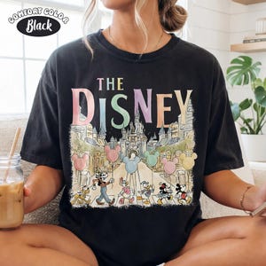 Pu&ograve; includere: T-shirt nero con "THE DISNEY" in colori pastello. La grafica presenta i personaggi Disney che attraversano una strada con palloncini di Topolino e un castello sullo sfondo. La maglietta ha un'etichetta Comfort Colors.