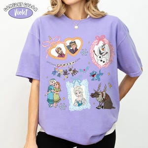 May include: A violet Comfort Colors t-shirt featuring various characters and elements from the animated film "Frozen." The design includes framed images of characters, snowflakes, and the word "FROZEN" in a banner.