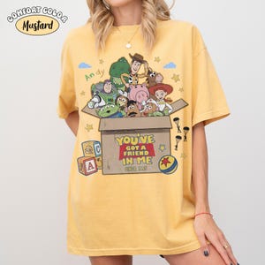 K&ouml;nnte beinhalten: Senffarbenes T-Shirt mit einer Grafik von Charakteren aus dem Film Toy Story. Das Design zeigt eine Box mit dem Text "YOU'VE GOT A FRIEND IN ME SINCE 1995" und dem Namen "Andy". Das Shirt hat ein Comfort Colors-Label.