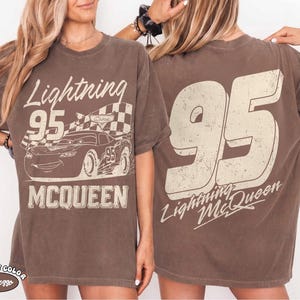 May include: Brown t-shirt featuring a graphic of Lightning McQueen from the Cars movie. The front displays the text "Lightning 95 McQueen" with a checkered flag and car illustration. The back shows a large "95" and "Lightning McQueen".