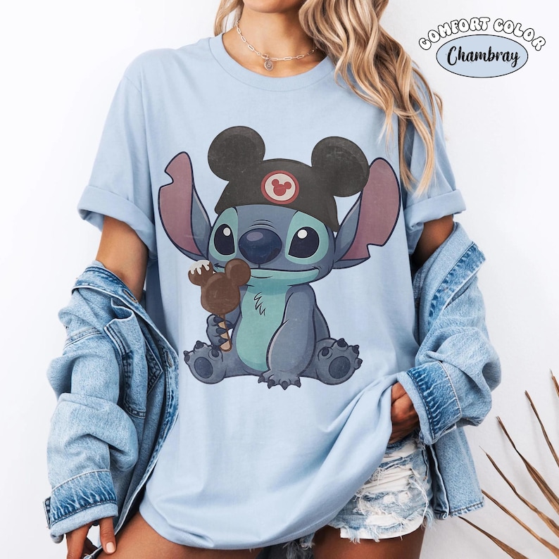 May include: A light blue t-shirt featuring a graphic of the character Stitch wearing black mouse ears and holding a brown chocolate treat. The text COMFORT COLOR Chambray is printed in the upper right corner of the image in a circular design.
