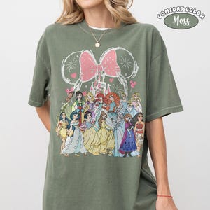Op de afbeelding: Olijfgroen T-shirt met een afbeelding van Disney-prinsessen. Het ontwerp bevat een roze strik, vuurwerk en een kasteel. Het shirt is gelabeld "Comfort Color Moss".