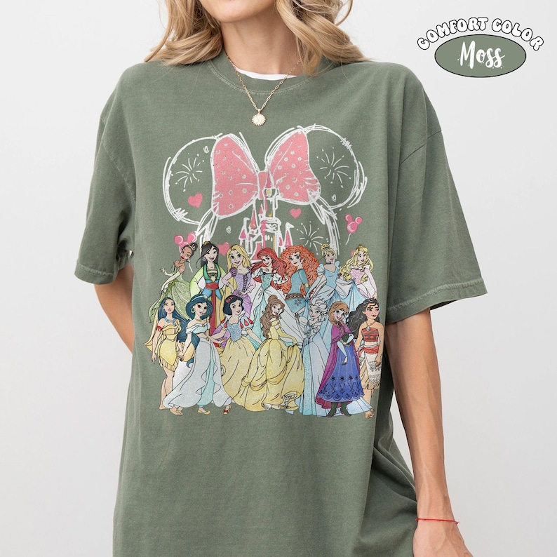 Op de afbeelding: Olijfgroen T-shirt met een afbeelding van Disney-prinsessen. Het ontwerp bevat een roze strik, vuurwerk en een kasteel. Het shirt is gelabeld "Comfort Color Moss".