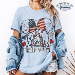 May include: Light blue t-shirt with a graphic featuring a castle, hearts, fireworks, and a bow in the American flag design. The shirt has rolled-up sleeves and the words "Comfort Color Chambray" are printed on the top right.