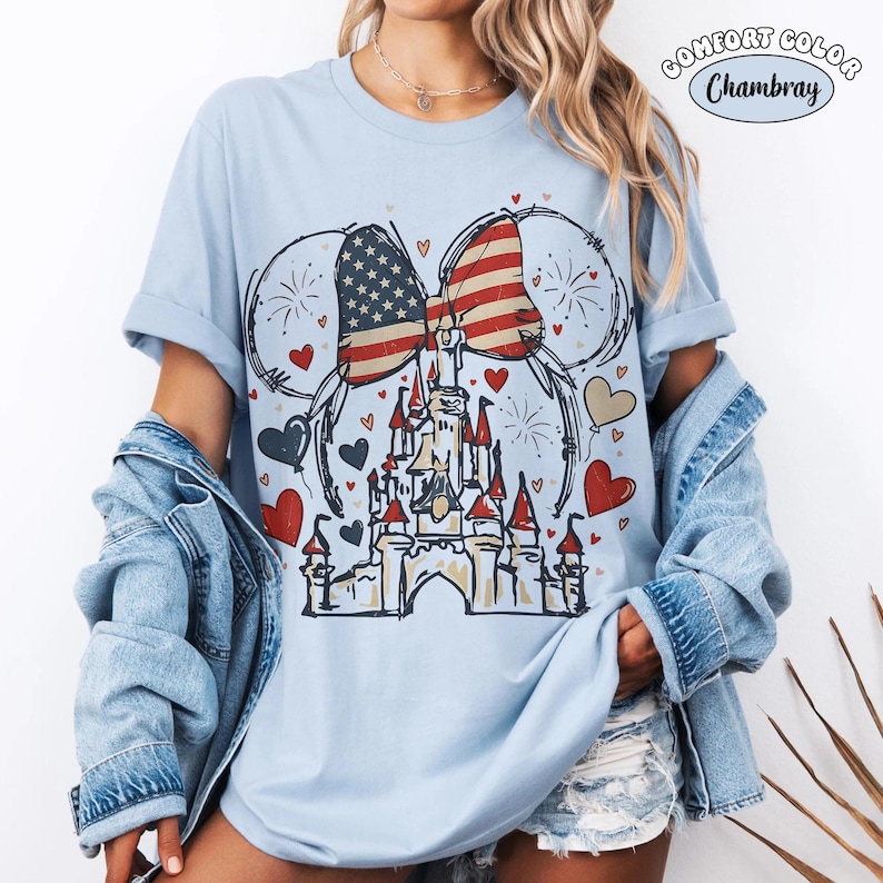 May include: Light blue t-shirt with a graphic featuring a castle, hearts, fireworks, and a bow in the American flag design. The shirt has rolled-up sleeves and the words "Comfort Color Chambray" are printed on the top right.