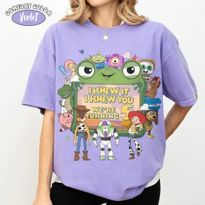 Puede incluir: Una camiseta de color violeta claro con un gr&aacute;fico de varios personajes animados que rodean un marco verde con forma de rana. El texto dentro del marco dice I KNEW IT I KNEW YOU WE'RE TURNING 5. Los personajes incluyen un vaquero, un guardi&aacute;n espacial, una vaquera, un cerdo y un dinosaurio.