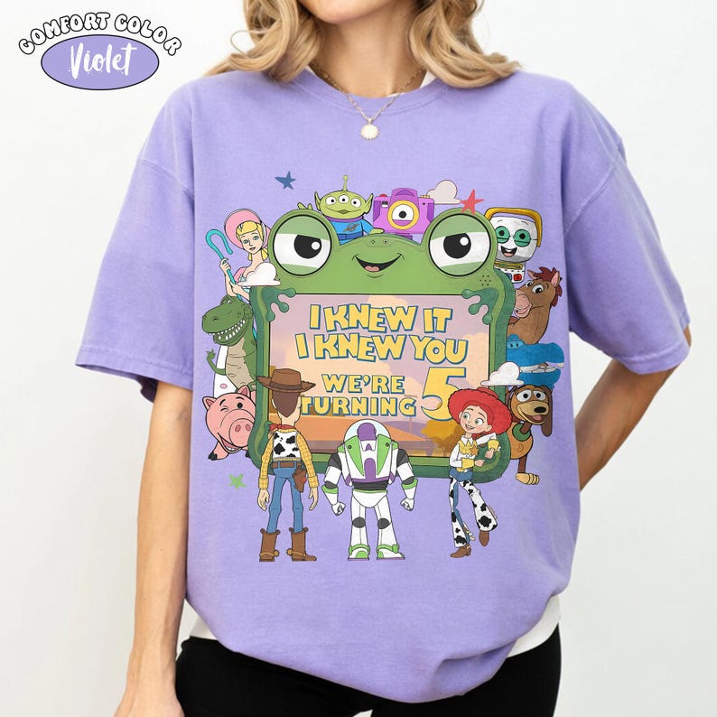 Puede incluir: Una camiseta de color violeta claro con un gr&aacute;fico de varios personajes animados que rodean un marco verde con forma de rana. El texto dentro del marco dice I KNEW IT I KNEW YOU WE'RE TURNING 5. Los personajes incluyen un vaquero, un guardi&aacute;n espacial, una vaquera, un cerdo y un dinosaurio.