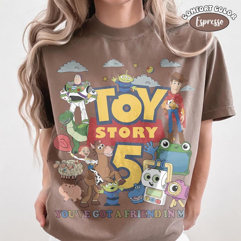 May include: An espresso brown short-sleeve t-shirt featuring a graphic print of Toy Story characters including Buzz Lightyear, Woody, Jessie, Bullseye, and others. The text reads Toy Story 5 and You've Got A Friend In Me. A Comfort Color label is visible.