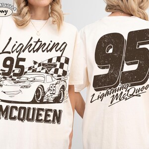 May include: Ivory-colored t-shirt featuring a distressed graphic of Lightning McQueen race car with the number 95 and the text "Lightning McQueen". The back of the shirt displays a large number 95 and "Lightning McQueen".