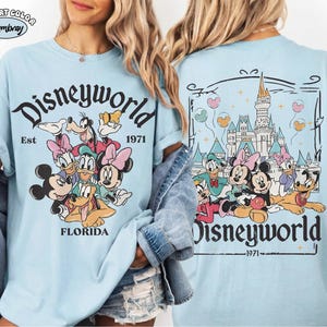 K&ouml;nnte beinhalten: Ein hellblaues T-Shirt mit einer Grafik von Disney-Figuren wie Mickey, Minnie, Donald, Daisy, Goofy und Pluto. Die Vorderseite zeigt den Text Disneyworld Est 1971 Florida. Die R&uuml;ckseite zeigt das Schloss mit dem Text Disneyworld 1971.