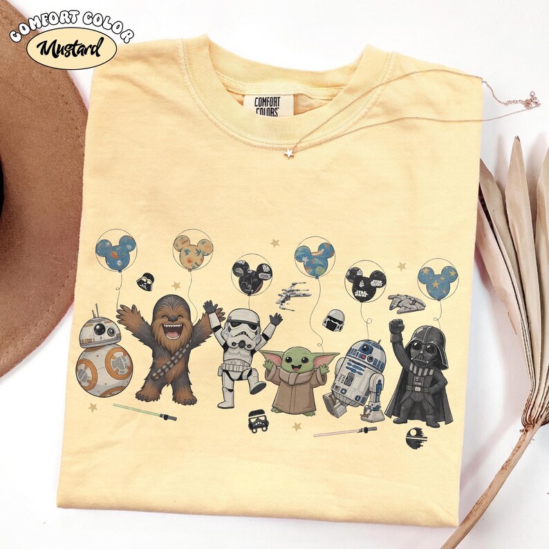 May include: Mustard-colored t-shirt featuring cartoon characters from Star Wars and Mickey Mouse balloons. The shirt has a crew neck and short sleeves. The text "Comfort Colors" is printed on the tag.