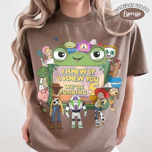 Puede incluir: Una camiseta color marr&oacute;n espresso con un gr&aacute;fico grande de un marco de rana verde que rodea varios personajes animados. El texto dentro del marco dice I KNEW IT I KNEW YOU WE'RE TURNING 5. El dise&ntilde;o incluye un vaquero y un guardi&aacute;n espacial.