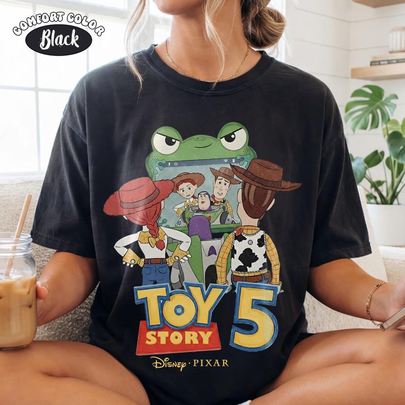 Pu&ograve; includere: T-shirt nera Comfort Colors con grafica di Toy Story. Il design presenta personaggi del film, il testo "Toy Story 5" e il logo Disney Pixar. Un personaggio verde a forma di rana &egrave; sullo sfondo.