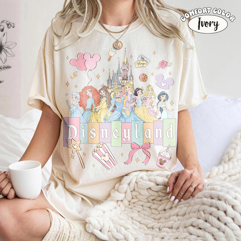 Puede incluir: Camiseta de color marfil con un dise&ntilde;o de Disneyland que presenta princesas de dibujos animados, un castillo y otros elementos tem&aacute;ticos. La camiseta tiene cuello redondo y mangas cortas. El texto "Disneyland" est&aacute; en colores pastel.