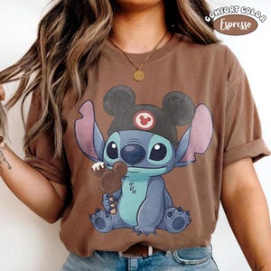 May include: An espresso brown short-sleeve t-shirt featuring a graphic of the character Stitch wearing a black hat with mouse ears and holding a brown mouse-shaped ice cream bar. The text Comfort Color Espresso is printed in the top right corner.