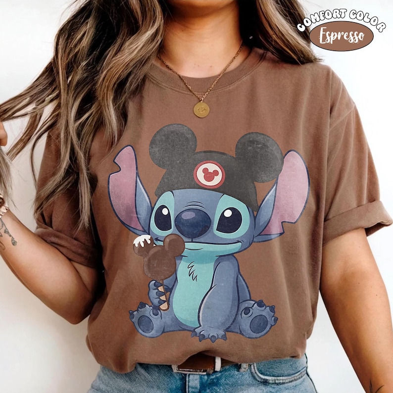 May include: An espresso brown short-sleeve t-shirt featuring a graphic of the character Stitch wearing a black hat with mouse ears and holding a brown mouse-shaped ice cream bar. The text Comfort Color Espresso is printed in the top right corner.