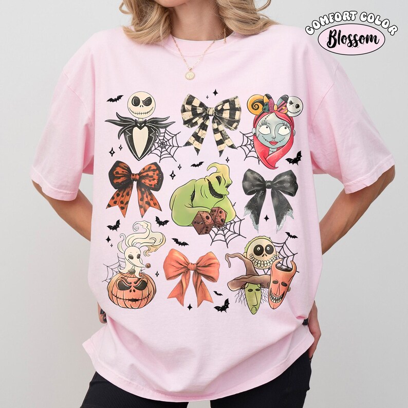 May include: A light pink t-shirt featuring a graphic print of characters and bows from The Nightmare Before Christmas, including Jack Skellington, Sally, Oogie Boogie, Zero, and Lock, Shock, and Barrel, with spiderwebs and bats throughout.