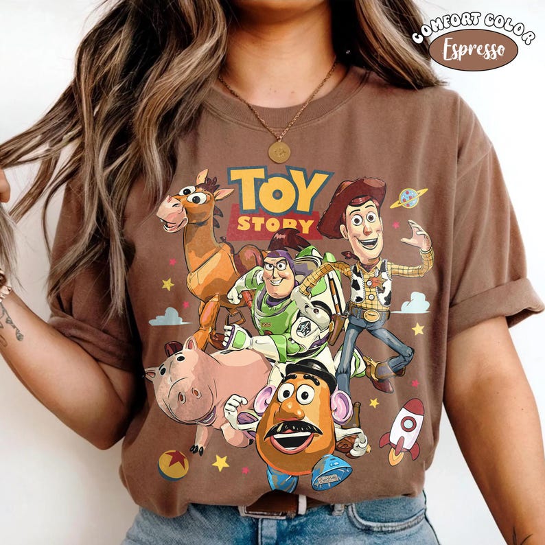 May include: Espresso-colored Comfort Colors t-shirt featuring a graphic of Toy Story characters, including Woody, Buzz Lightyear, and Mr. Potato Head. The text "TOY STORY" is displayed above the characters. The shirt has rolled-up sleeves.