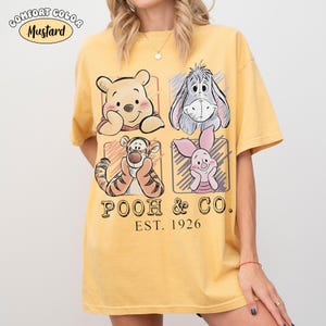 May include: Mustard yellow t-shirt featuring four cartoon character illustrations. The shirt includes Winnie the Pooh, Eeyore, Tigger, and Piglet, with the text "POOH & CO. EST. 1926". The shirt has a Comfort Color label.