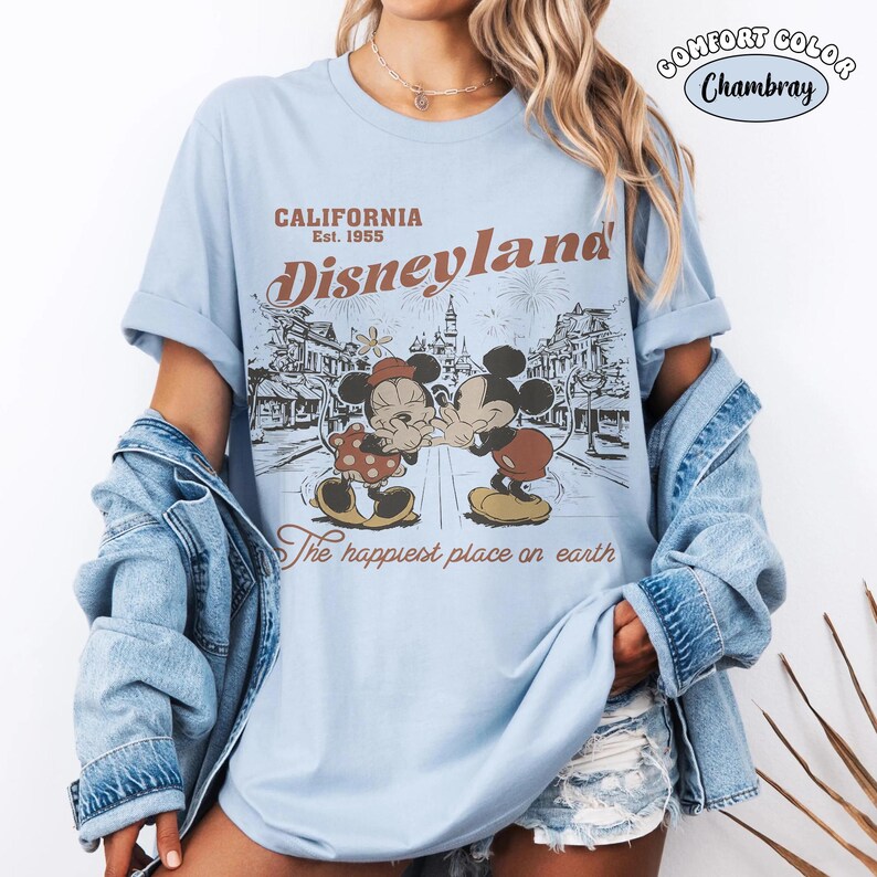 May include: A light blue t-shirt featuring a graphic print of Mickey and Minnie Mouse. The text reads California Est. 1955 Disneyland The happiest place on earth. A circular badge in the top right corner displays the text Comfort Color Chambray.