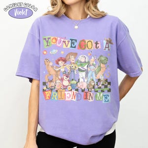 May include: A violet Comfort Colors t-shirt featuring a colorful graphic of characters from the movie Toy Story. The text "YOU'VE GOT A FRIEND IN ME" is displayed above the characters. The shirt has short sleeves and a crew neck.