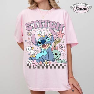 Puede incluir: Camiseta rosa claro con un dibujo animado de Stitch, patos y flores. La palabra "STITCH" est&aacute; encima del dibujo en una fuente de burbujas. La camiseta tiene una etiqueta Comfort Color Blossom.