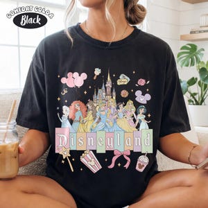Puede incluir: Camiseta negra Comfort Colors con un gr&aacute;fico colorido de Disneyland. El dise&ntilde;o incluye princesas de Disney, un castillo y la palabra "Disneyland" en letras pastel. Gr&aacute;ficos adicionales incluyen iconos de Mickey Mouse, palomitas de ma&iacute;z y una varita m&aacute;gica.