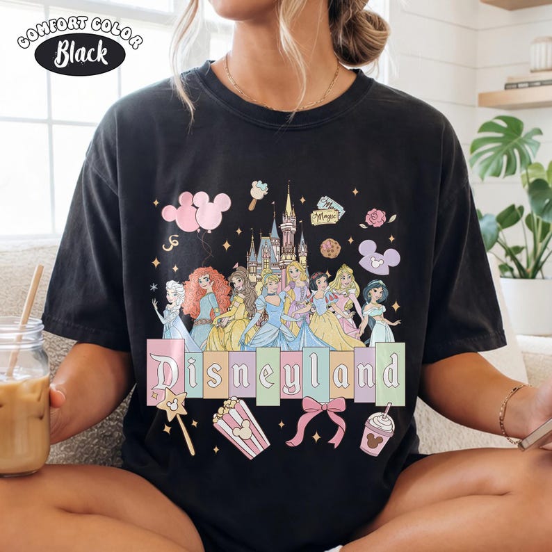 Puede incluir: Camiseta negra Comfort Colors con un gr&aacute;fico colorido de Disneyland. El dise&ntilde;o incluye princesas de Disney, un castillo y la palabra "Disneyland" en letras pastel. Gr&aacute;ficos adicionales incluyen iconos de Mickey Mouse, palomitas de ma&iacute;z y una varita m&aacute;gica.