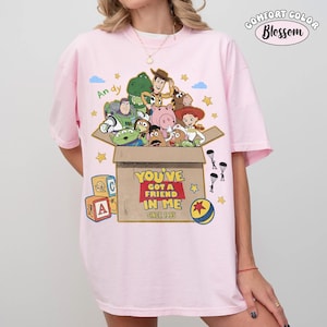 K&ouml;nnte beinhalten: Hellrosa T-Shirt mit einer Grafik von Toy Story-Charakteren in einer Pappschachtel. Der Text "YOU'VE GOT A FRIEND IN ME SINCE 1995" ist auf der Box aufgedruckt. Das Shirt hat auch den Text "Andy" und das Comfort Color Blossom Logo.