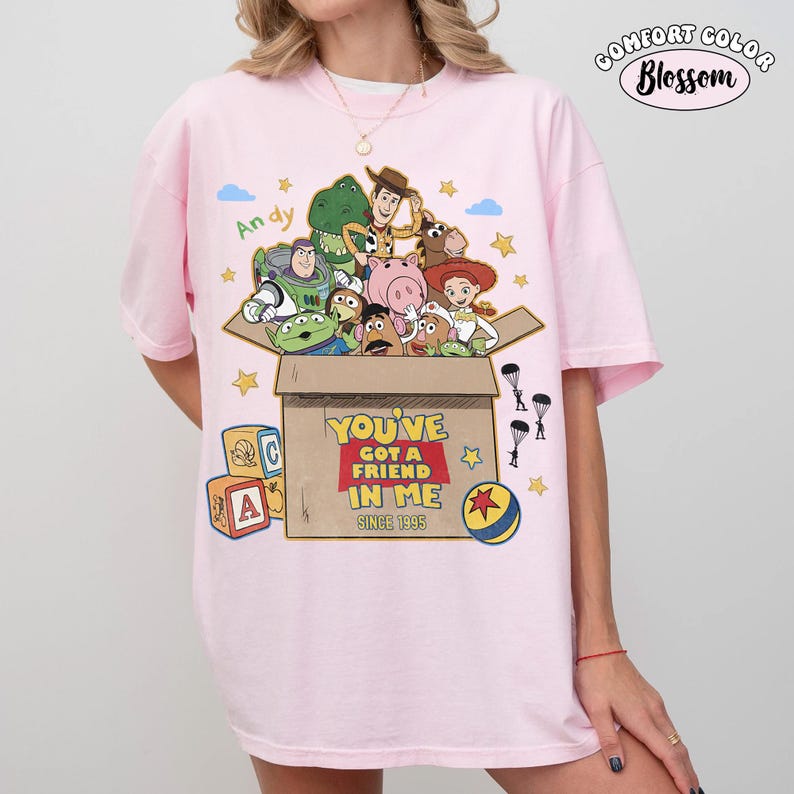 K&ouml;nnte beinhalten: Hellrosa T-Shirt mit einer Grafik von Toy Story-Charakteren in einer Pappschachtel. Der Text "YOU'VE GOT A FRIEND IN ME SINCE 1995" ist auf der Box aufgedruckt. Das Shirt hat auch den Text "Andy" und das Comfort Color Blossom Logo.