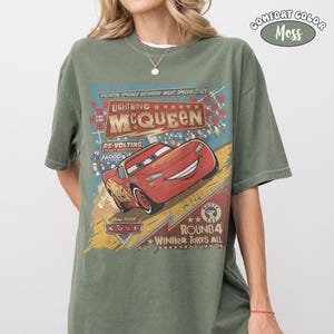 May include: Olive green t-shirt featuring a graphic of Lightning McQueen from the movie Cars. The design includes text that reads "Lightning McQueen," "Radiator Springs," and "Round 4 Winner Takes All." The shirt is labeled "Comfort Color Moss."