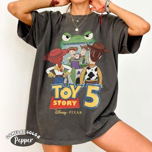 Pu&ograve; includere: T-shirt Comfort Colors grigio scuro con grafica di Toy Story 5. Il design presenta personaggi come Woody, Buzz Lightyear e Jessie, insieme al logo Toy Story e al marchio Disney Pixar. La maglietta ha un aspetto vintage e usurato.
