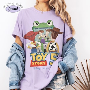 Pu&ograve; includere: Una maglietta lilla Comfort Colors con una grafica del film Disney Pixar Toy Story 5. Il design include personaggi come Woody, Buzz Lightyear e Jessie. La maglietta presenta la scritta "TOY STORY" e il logo Disney Pixar.