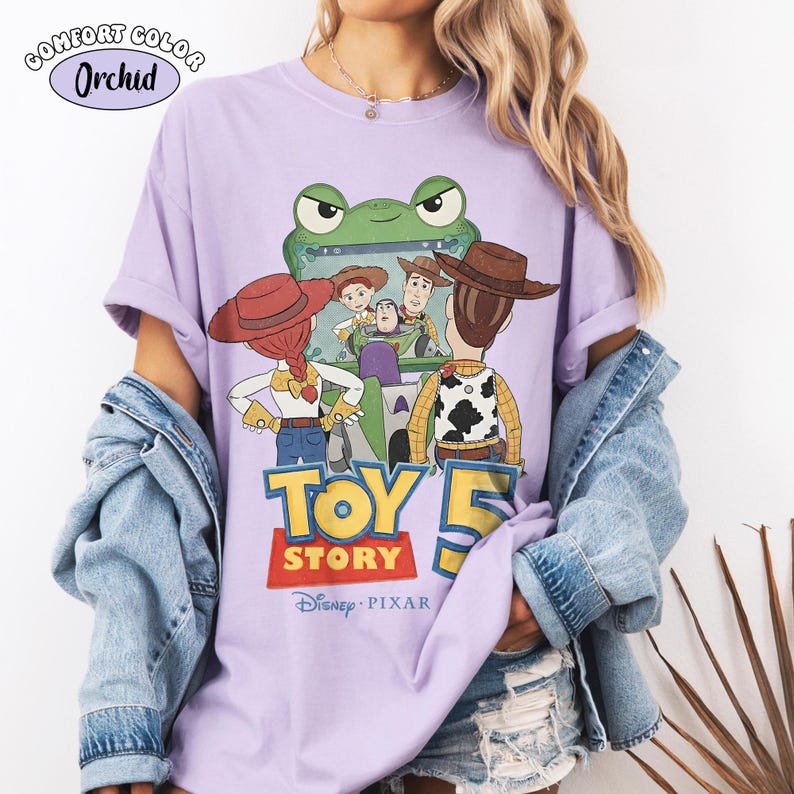 Pu&ograve; includere: Una maglietta lilla Comfort Colors con una grafica del film Disney Pixar Toy Story 5. Il design include personaggi come Woody, Buzz Lightyear e Jessie. La maglietta presenta la scritta "TOY STORY" e il logo Disney Pixar.