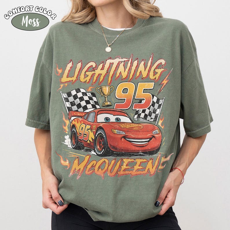 Puede incluir: Una camiseta verde musgo con un gr&aacute;fico de un coche de carreras rojo con el n&uacute;mero 95, un trofeo dorado y banderas a cuadros. El texto dice LIGHTNING 95 MCQUEEN en una fuente estilizada, rodeado de llamas naranjas y una etiqueta Comfort Color Moss.
