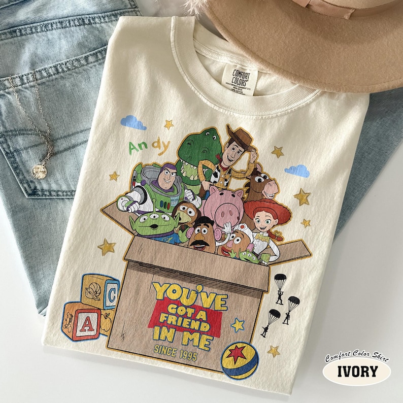 K&ouml;nnte beinhalten: Elfenbeinfarbenes Comfort Colors T-Shirt mit einer Grafik von Toy Story-Charakteren in einer Box. Der Text "YOU'VE GOT A FRIEND IN ME SINCE 1995" ist darunter gedruckt. Das Shirt liegt auf einer Jeans.