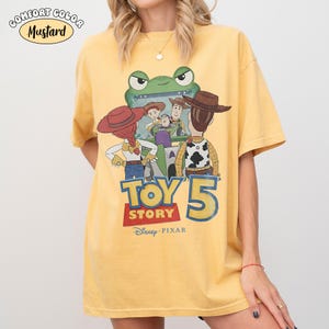 Pu&ograve; includere: T-shirt color senape con una grafica dei personaggi del film "Toy Story 5". Il design include Woody, Buzz Lightyear e altri personaggi. Il testo "TOY STORY 5" &egrave; visualizzato sotto i personaggi, con il logo Disney Pixar.