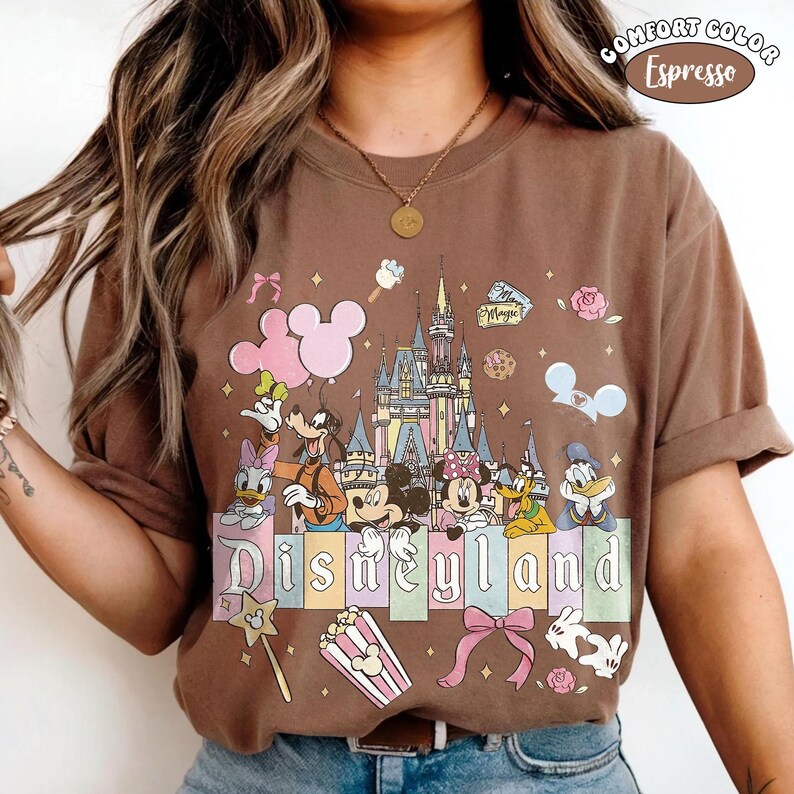 Peut inclure: T-shirt Comfort Colors couleur expresso avec un motif Disneyland. Le design comprend Mickey et Minnie Mouse, Donald Duck, Dingo et Daisy Duck, un ch&acirc;teau, du pop-corn et le mot "Disneyland".