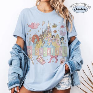 Puede incluir: Camiseta azul claro con un gr&aacute;fico de Disneyland que presenta princesas de Disney, un castillo e iconos tem&aacute;ticos. La palabra "Disneyland" est&aacute; impresa en colores pastel. La camiseta es de material Comfort Colors Chambray.