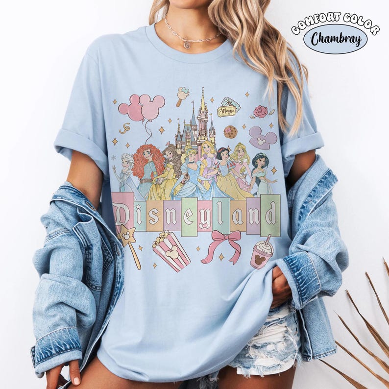 Puede incluir: Camiseta azul claro con un gr&aacute;fico de Disneyland que presenta princesas de Disney, un castillo e iconos tem&aacute;ticos. La palabra "Disneyland" est&aacute; impresa en colores pastel. La camiseta es de material Comfort Colors Chambray.