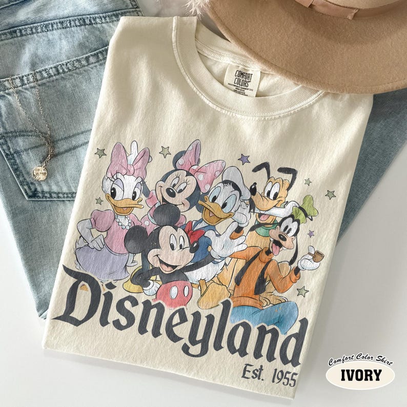 Pu&ograve; includere: Una t-shirt a girocollo color avorio con una stampa grafica di Mickey Mouse, Minnie Mouse, Donald Duck, Daisy Duck, Goofy e Pluto. Il testo Disneyland Est. 1955 &egrave; stampato sotto i personaggi con un carattere scuro. Etichetta Comfort Colors.