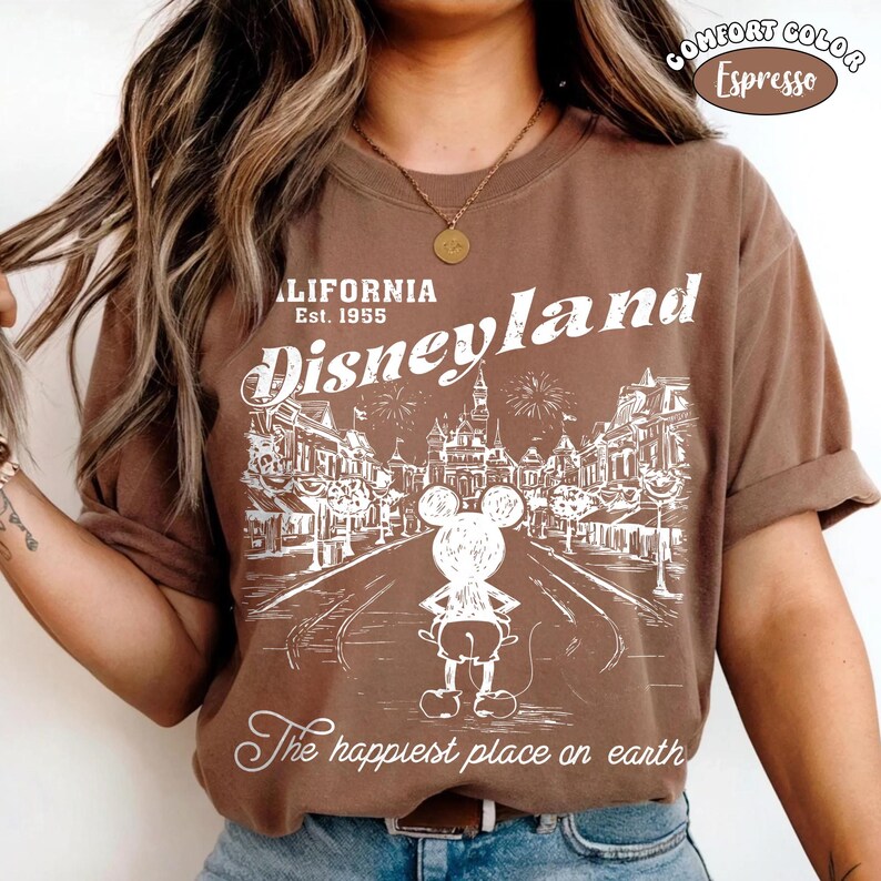 May include: Espresso-colored t-shirt with a white graphic of Disneyland. The design includes the words "California Disneyland Est. 1955" and "The happiest place on earth" with an illustration of Mickey Mouse and the park.