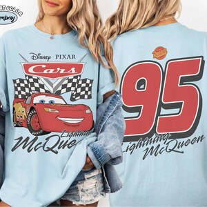 Może przedstawiać: Jasnoniebieska koszulka z Lightning McQueen z filmu Auta Disneya Pixara. Prz&oacute;d przedstawia postać z napisem "Cars" i "Lightning McQueen". Tył pokazuje duży napis "95" i tekst "Lightning McQueen".