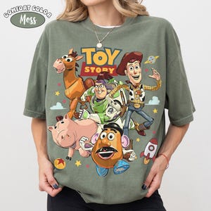 May include: A moss green Comfort Colors t-shirt featuring a colorful graphic of Toy Story characters. The design includes Woody, Buzz Lightyear, and other characters from the animated film. The text "TOY STORY" is in yellow and red.