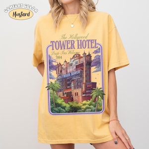 Op de afbeelding: Mosterdkleurig T-shirt met een afbeelding van het Hollywood Tower Hotel. Het ontwerp bevat de tekst "The Hollywood Tower Hotel" en "Drop-Ins Welcome 1994" in een paarse en witte lijst. Palmbomen zijn ook in het ontwerp opgenomen.