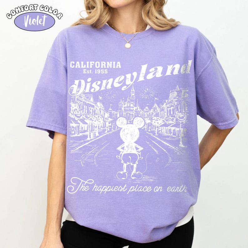 May include: Violet Comfort Colors t-shirt featuring a Disneyland design. The design includes the text "CALIFORNIA Est. 1955 Disneyland" and a graphic of Mickey Mouse. The phrase "The happiest place on earth" is also included.