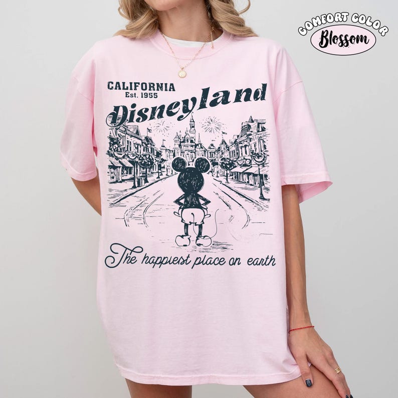 May include: Pink t-shirt with a Disneyland graphic. The design features a black and white illustration of Mickey Mouse in front of a street scene with the text "California Est. 1955 Disneyland" and "The happiest place on earth."