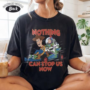 May include: Black Comfort Colors t-shirt featuring Woody and Buzz Lightyear from Toy Story. The graphic shows the characters in a race car with the text "NOTHING CAN STOP US NOW" in red. The shirt has a vintage look.
